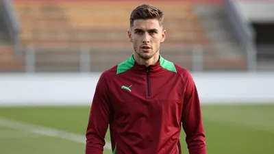 A determined 22-year-old right-back with a lean build, wearing a plain dark red and green training top, stands on the...