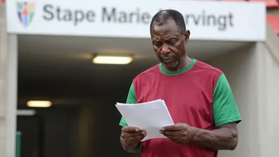 A 50-year-old, ruggedly-built, African male football coach in his late 40s, with a weathered face and a determined...