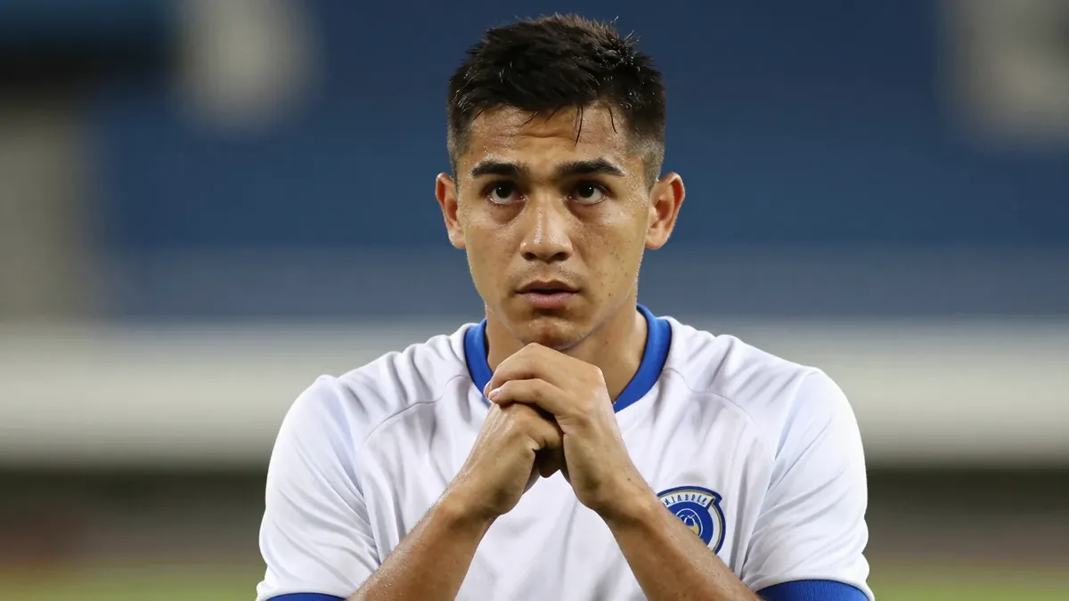 A young coach in a clean white and blue jersey, head-and-shoulders, wearing a thoughtful expression, his hands clasped...