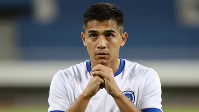 A young coach in a clean white and blue jersey, head-and-shoulders, wearing a thoughtful expression, his hands clasped...