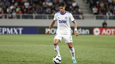 A 25-year-old Le Mans FC midfielder in a white jersey with a blue stripe, playing with a ball at his feet, on a green...