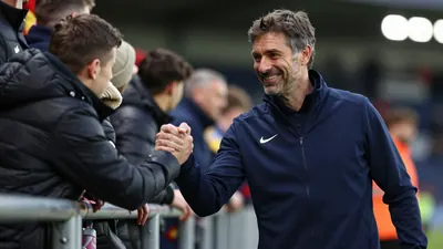 A 40-year-old football manager in a dark blue tracksuit, standing close to the touchline, smiling warmly as he shakes...