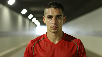 Mid-30s lean Le Mans FC forward, intense gaze, dark red jersey with subtle green piping, sweat-glazed skin, hair...