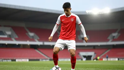 A 20-year-old Le Mans FC midfielder in a floodlit stadium, wearing a red jersey with white sleeves, and white shorts...