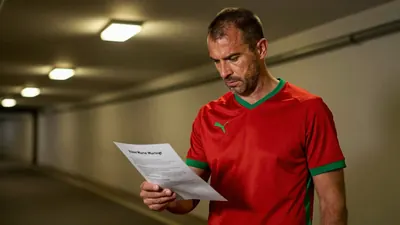 A 35-year-old, 5'10" male coach with a rugged build, wearing a plain, dark red and green polyester home kit, stands in...