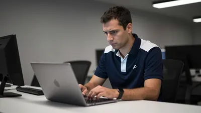 A concerned Le Mans FC staff member in a navy blue and white kit, mid-30s, with short dark hair, reviewing a laptop...